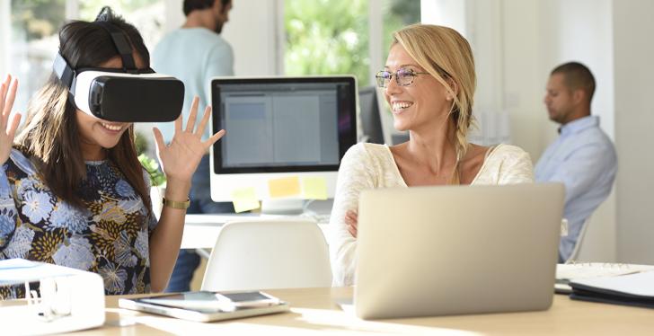 Zwei Frauen an einem Schreibtisch; eine von ihnen trägt eine VR-Brille...
