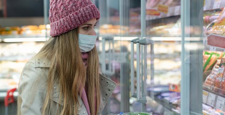 Eine Frau mit Mütze, Mundschutz und Handschuhen vor einem Kühlregal im...