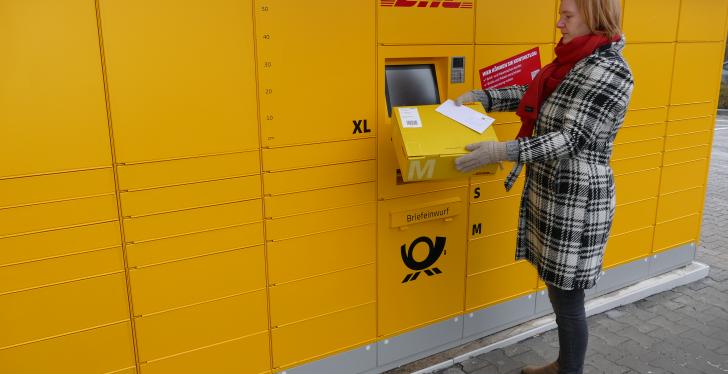 Frau gibt Paket an gelber Poststation ab