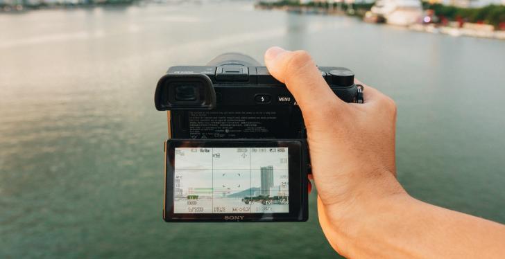 Hand hält eine Kamera und hält diese über einen Fluss, mit einer Stadt im...