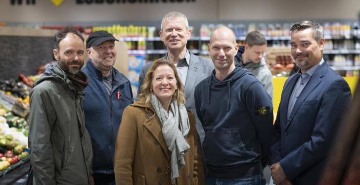 6 Personen stehen in einem Supermarkt für ein Gruppen-Foto zusammen....
