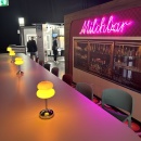 Barbereich mit gelben Tischlampen und pinkem Neon-Schriftzug „Milchbar“....