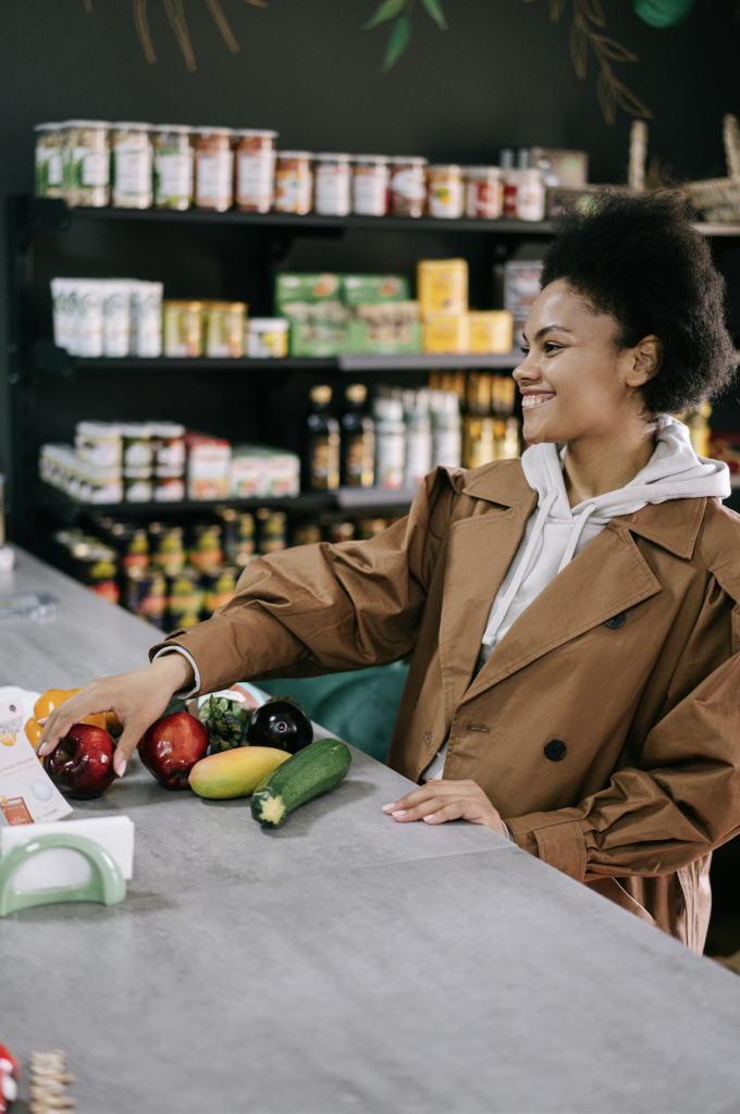 A woman in a brown coat smiles while selecting fresh vegetables and fruits at a...
