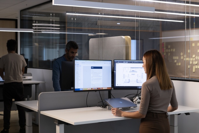 Frau seht am Schreibtisch, auf dem 2 Bildschirme stehen...