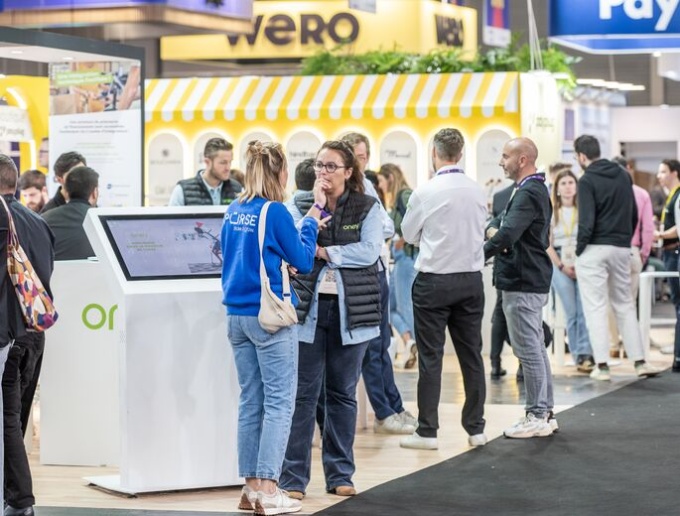 Menschen stehen in einer Messehalle vor einem Messestand und unterhalten sich...