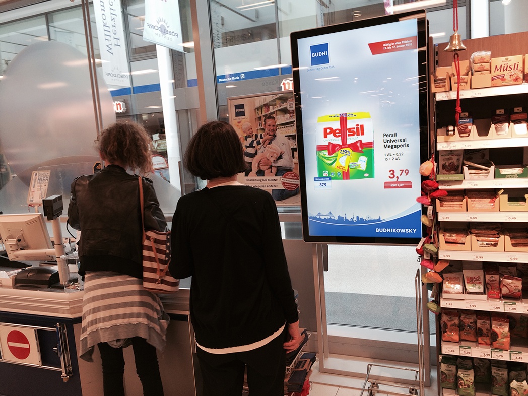Foto: Werbebildschirm in einem Drogeriemarkt an der Kasse mit wartenden Kunden;...