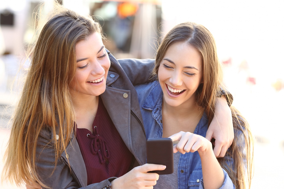 Zwei Mädchen umarmen sich lächelnd und schauen auf ein Smartphone....