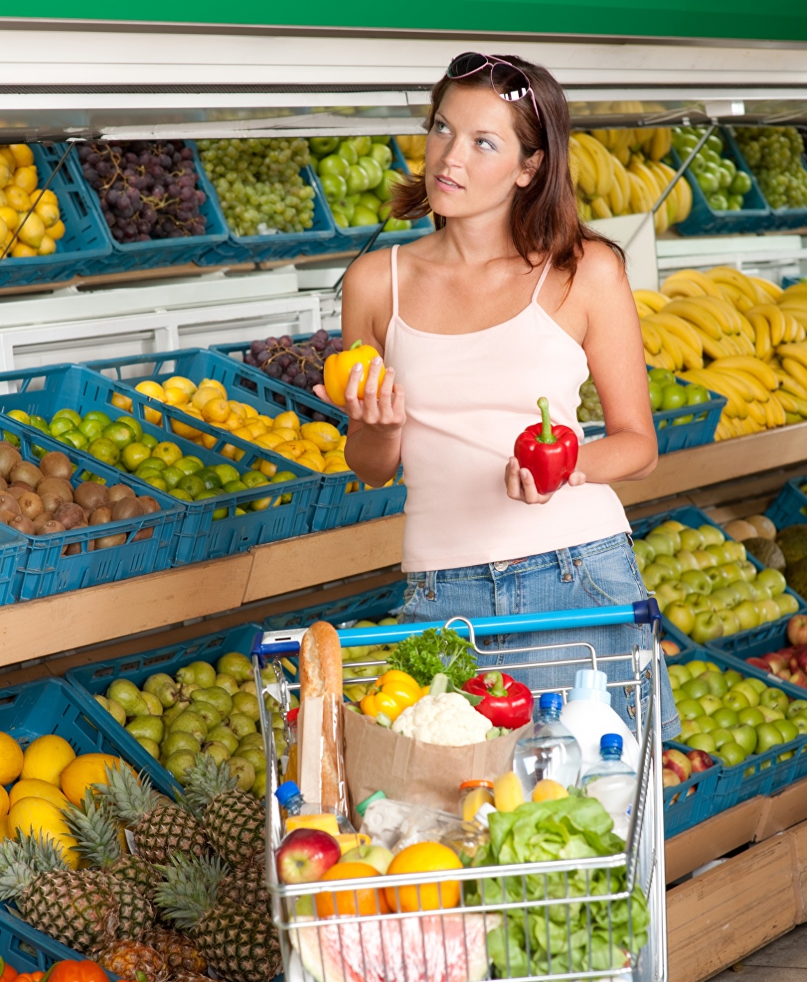 Frau mit Einkaufswagen im Supermarkt hält rote und gelbe Paprika in der Hand;...