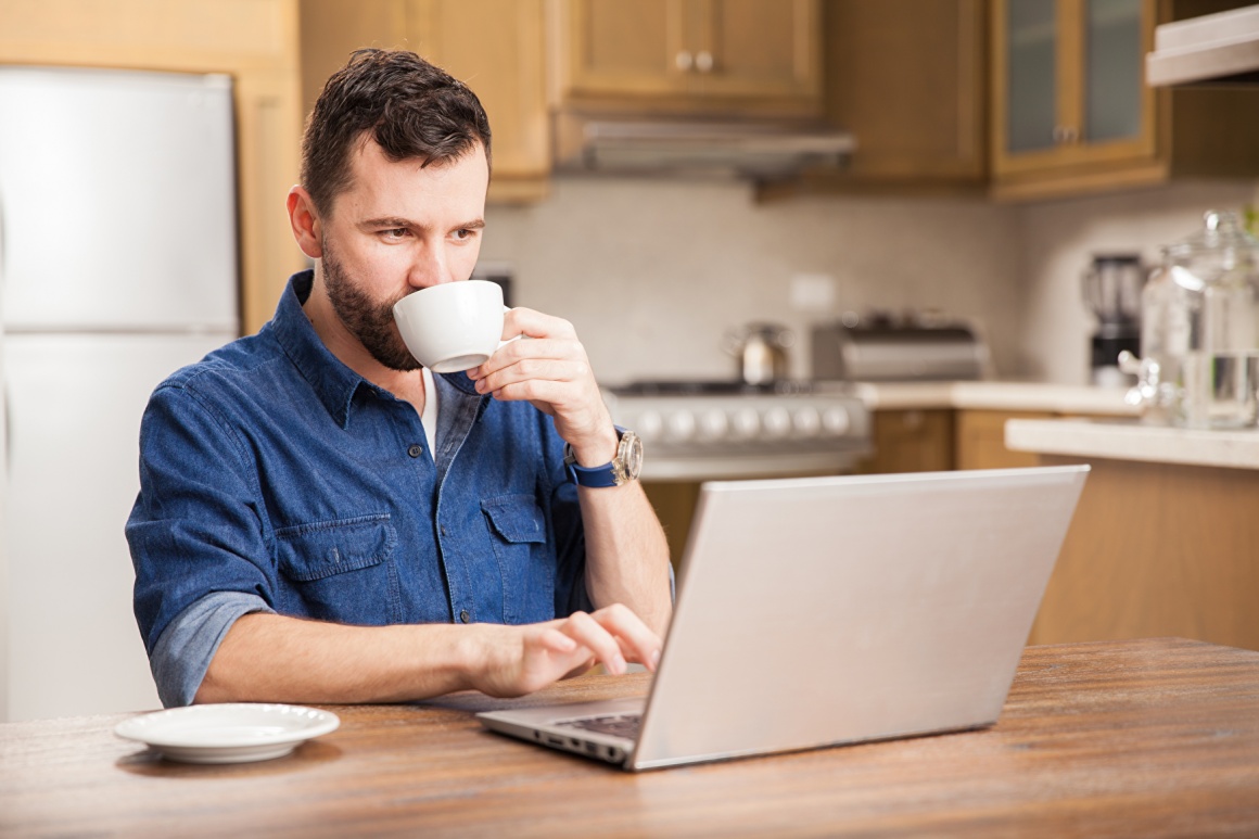 Mann sitzt mit Laptop und Kaffee am Küchentisch und arbeitet...